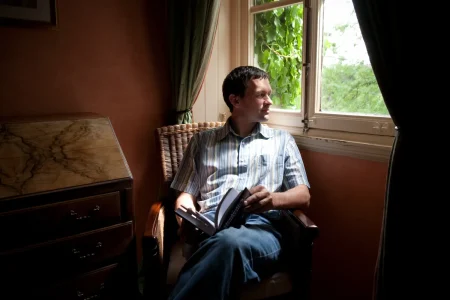 Man sitting in chair with book open in his hands as he stares out the window thinking about addiction
