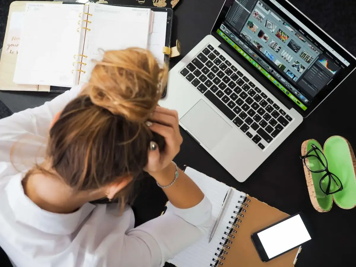 Photograph taken from above of a woman working from home with alcohol addiction, holding her head in her hands as she sits in front of a laptop with a diary, a journal, and a phone all around her
