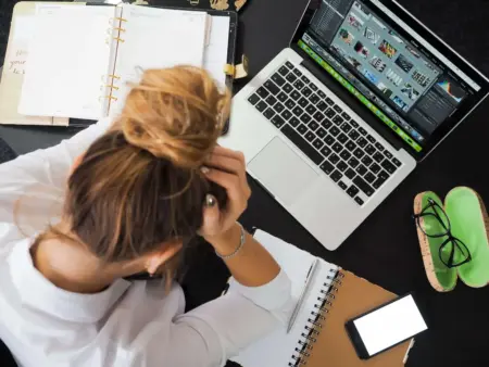 Photograph taken from above of a woman working from home with alcohol addiction, holding her head in her hands as she sits in front of a laptop with a diary, a journal, and a phone all around her