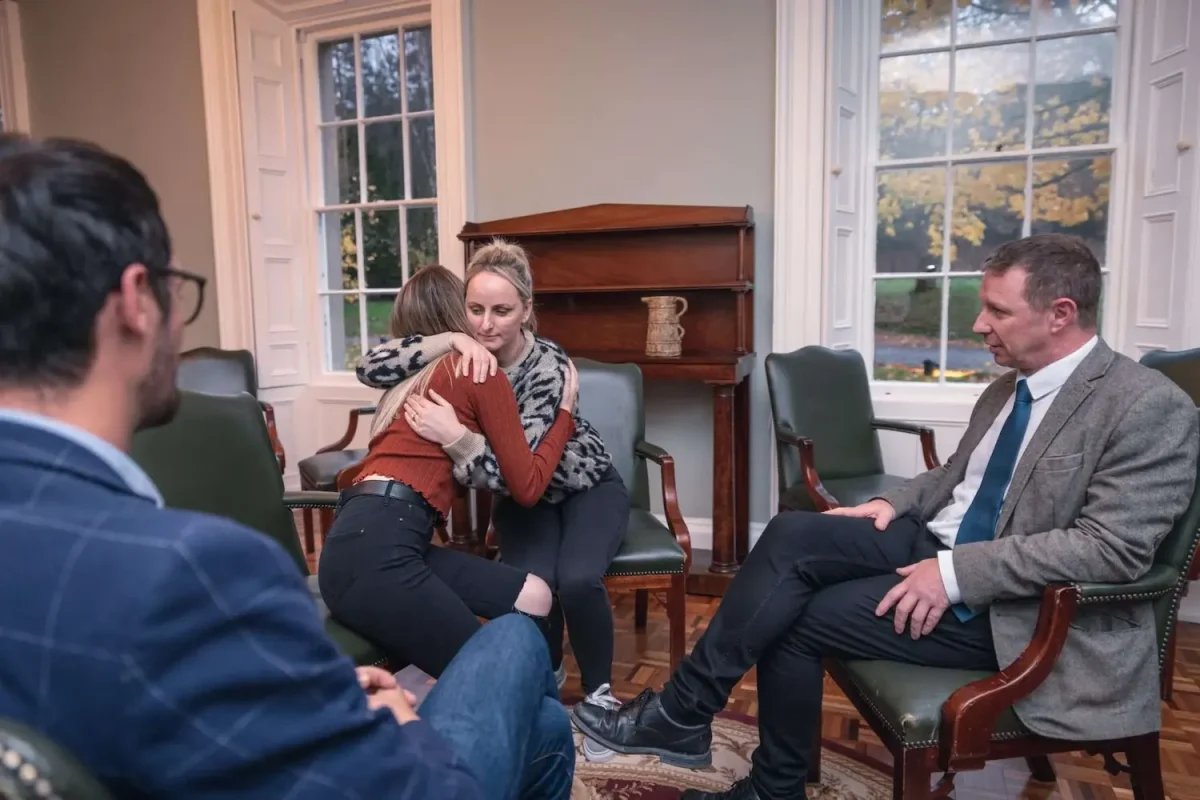 Picture depicting family therapy where four people sitting in a large room in a circle, two of them are embracing