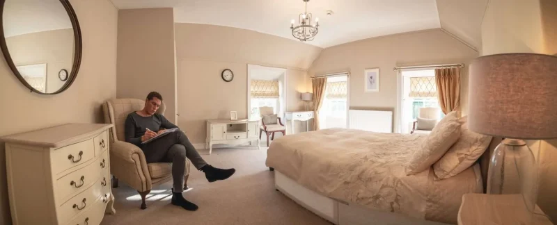 A patient doing reflective work as part of his rehab programme sitting in an armchair in the corner of his room which also contains a double bed, dresser, chairs, desks, and several windows letting lots of natural light in