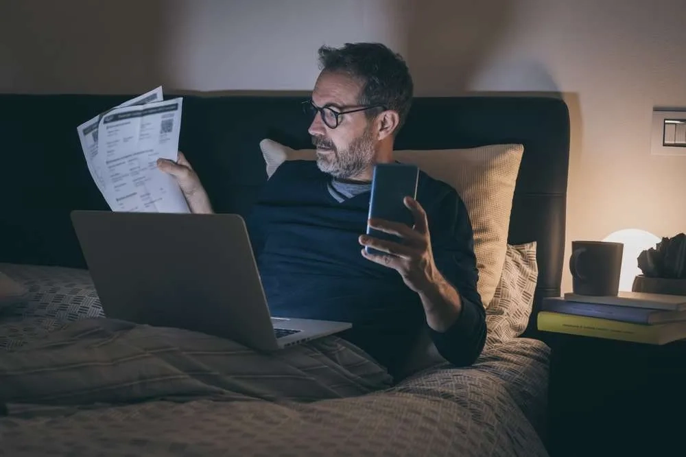 Image depicting work addiction of a man sitting on his bed, with the light from his laptop screen lighting up his face while he looks at paperwork, and his phone