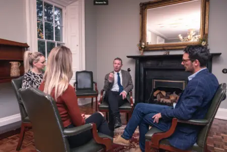 Four people sitting in a circle in front of a large fire place at Smarmore Castle discussing staging an intervention.