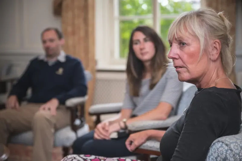 Photo of a woman in focus with others out of focus, all sitting in chairs in a circle during addiction rehab group therapy