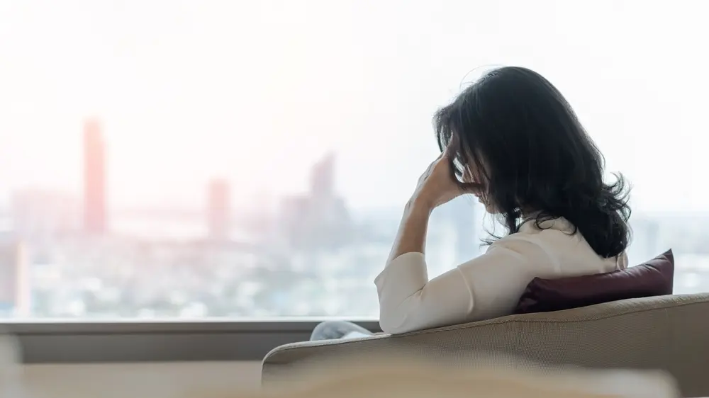 A woman sitting with her left hand holding her head, feeling anxious about the effects of menopause and addiction.